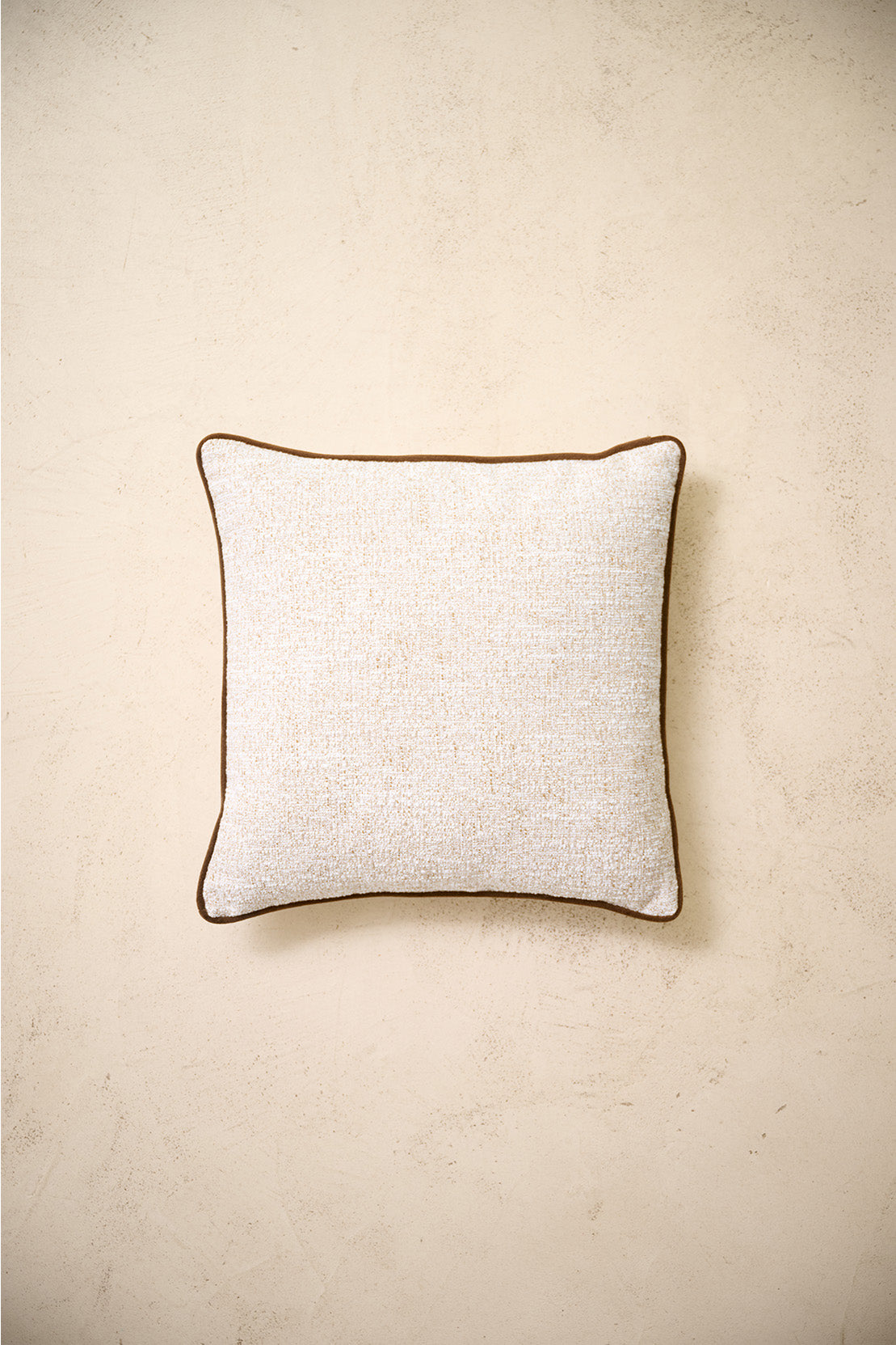 coussin carré CARIBOU en bouclé écru à passepoil marron, sur un mur en enduit crème