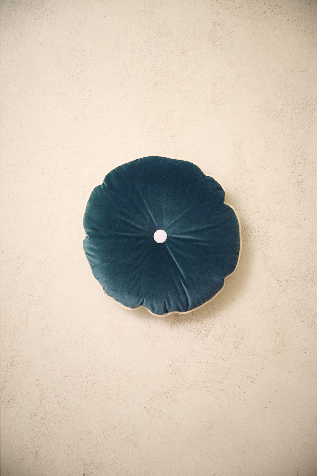 coussin rond MOON en velours bleu canard à bouton écru, posé sur un mur en enduit crème
