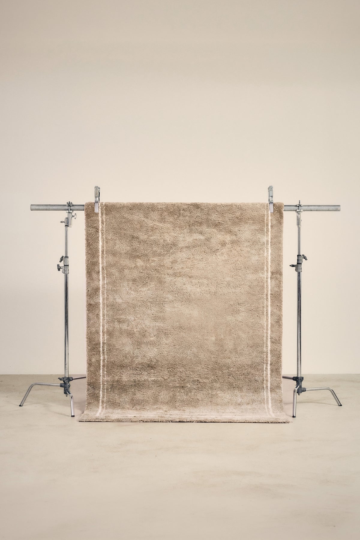 tapis ESTERO beige uni bordé de deux liserés écrus, suspendu sur un portant de studio