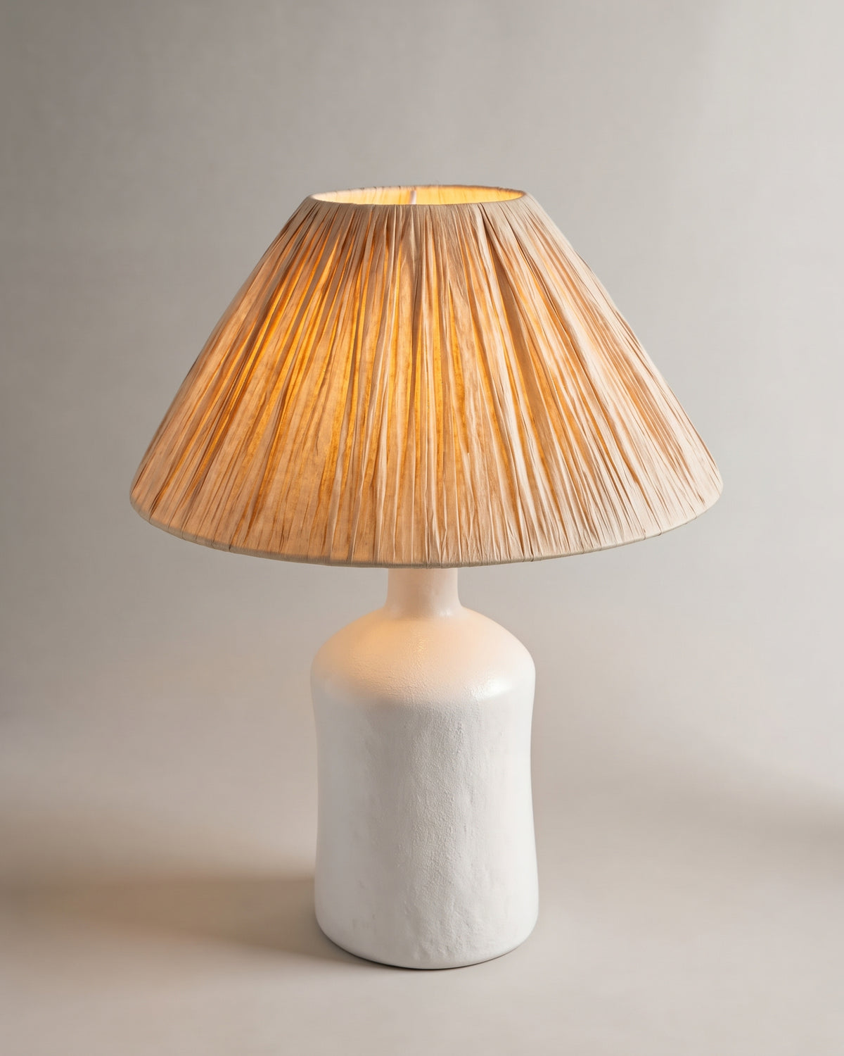 lampe de table KATARINA allumée, pied bouteille en céramique blanche et abat-jour en raphia plissé